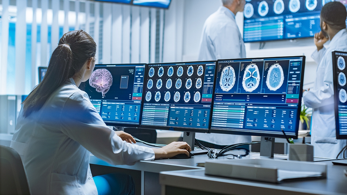 Frau in weißem Kittel vor einer Reihe von Monitoren die einen CT Scan eines Gehirns zeigen